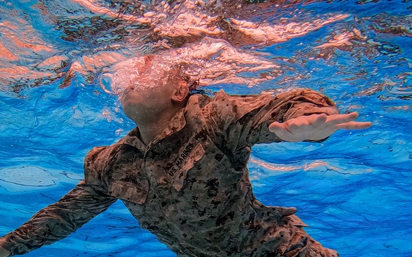 An underwater photograph taken inside a swimming pool shows a U.S. Marine in full camouflage uniform (MARPAT pattern) swimming beneath the surface. The image is captured from below, providing a unique perspective looking up toward the water surface. Additional personnel in uniform are visible in the background at the pool bottom, suggesting a military water survival or combat swimmer training exercise.