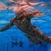 An underwater photograph taken inside a swimming pool shows a U.S. Marine in full camouflage uniform (MARPAT pattern) swimming beneath the surface. The image is captured from below, providing a unique perspective looking up toward the water surface. Additional personnel in uniform are visible in the background at the pool bottom, suggesting a military water survival or combat swimmer training exercise.