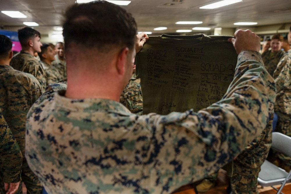 A U.S. Marine in MARPAT camouflage uniform holds up an olive drab garment (likely a t-shirt or bag) with squad rosters written in marker, displaying names organized under Squad 1, Squad 2, and Squad 3 headings. The setting appears to be an indoor military space, possibly aboard a ship or in a barracks, with multiple other Marines visible in the background. This appears to be a briefing or organizational meeting where squad assignments are being announced or reviewed.