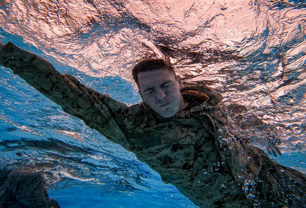 An underwater photograph captures a military service member swimming fully clothed in camouflage uniform. The image is taken from below the surface, showing the swimmer moving through the water with eyes closed and arms extended. A name tape reading 'NESTEB' (partially visible) is visible on the uniform, consistent with U.S. Marine Corps digital camouflage (MARPAT).
