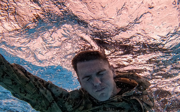 An underwater photograph captures a military service member swimming fully clothed in camouflage uniform. The image is taken from below the surface, showing the swimmer moving through the water with eyes closed and arms extended. A name tape reading 'NESTEB' (partially visible) is visible on the uniform, consistent with U.S. Marine Corps digital camouflage (MARPAT).