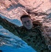 An underwater photograph captures a military service member swimming fully clothed in camouflage uniform. The image is taken from below the surface, showing the swimmer moving through the water with eyes closed and arms extended. A name tape reading 'NESTEB' (partially visible) is visible on the uniform, consistent with U.S. Marine Corps digital camouflage (MARPAT).