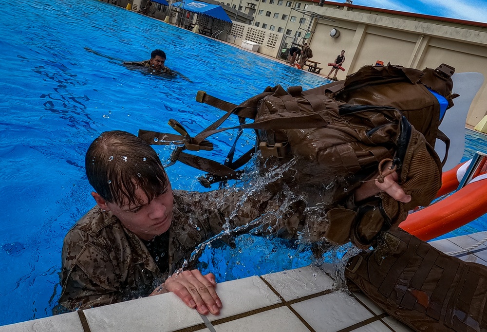Military personnel in full combat gear and camouflage uniforms are conducting water survival or combat swimming training in an outdoor pool facility. A soldier in the foreground is climbing out of the pool while wearing a heavy tactical backpack dripping with water, while another swimmer is visible in the background. The setting appears to be a military base with buildings and a blue canopy tent visible in the background.