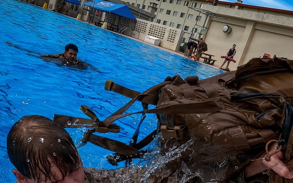 Military personnel in full combat gear and camouflage uniforms are conducting water survival or combat swimming training in an outdoor pool facility. A soldier in the foreground is climbing out of the pool while wearing a heavy tactical backpack dripping with water, while another swimmer is visible in the background. The setting appears to be a military base with buildings and a blue canopy tent visible in the background.