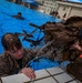 Military personnel in full combat gear and camouflage uniforms are conducting water survival or combat swimming training in an outdoor pool facility. A soldier in the foreground is climbing out of the pool while wearing a heavy tactical backpack dripping with water, while another swimmer is visible in the background. The setting appears to be a military base with buildings and a blue canopy tent visible in the background.