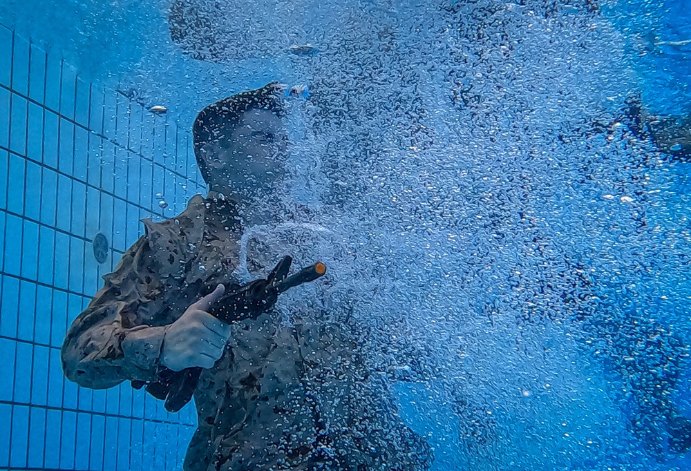 An underwater photograph taken inside a swimming pool showing a person in military-style camouflage uniform submerged and holding what appears to be a weapon or training device. A large cloud of bubbles surrounds the subject, likely from the discharge of the device or from the person exhaling. The tiled pool wall is visible in the background, suggesting this is a controlled training environment.