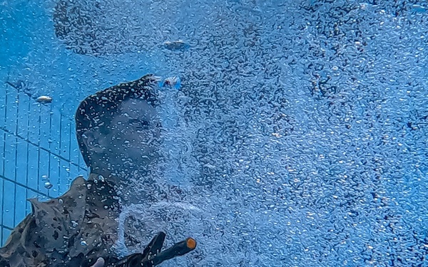 An underwater photograph taken inside a swimming pool showing a person in military-style camouflage uniform submerged and holding what appears to be a weapon or training device. A large cloud of bubbles surrounds the subject, likely from the discharge of the device or from the person exhaling. The tiled pool wall is visible in the background, suggesting this is a controlled training environment.
