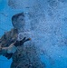 An underwater photograph taken inside a swimming pool showing a person in military-style camouflage uniform submerged and holding what appears to be a weapon or training device. A large cloud of bubbles surrounds the subject, likely from the discharge of the device or from the person exhaling. The tiled pool wall is visible in the background, suggesting this is a controlled training environment.