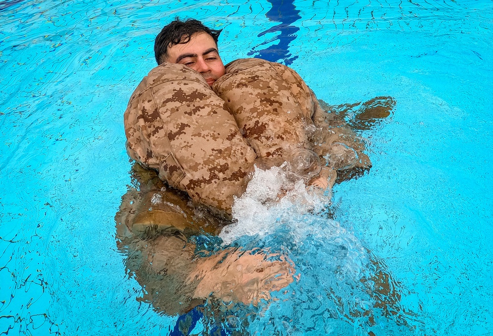 A person in military digital camouflage uniform is floating in a bright blue swimming pool, using an inflated camouflage flotation device wrapped around their torso. The individual appears to be undergoing military water survival training, keeping their head above water while kicking their legs. The image is taken from above at a slight angle, capturing the splashing water and the buoyancy aid in use.