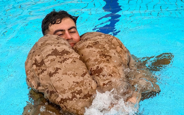 A person in military digital camouflage uniform is floating in a bright blue swimming pool, using an inflated camouflage flotation device wrapped around their torso. The individual appears to be undergoing military water survival training, keeping their head above water while kicking their legs. The image is taken from above at a slight angle, capturing the splashing water and the buoyancy aid in use.