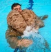 A person in military digital camouflage uniform is floating in a bright blue swimming pool, using an inflated camouflage flotation device wrapped around their torso. The individual appears to be undergoing military water survival training, keeping their head above water while kicking their legs. The image is taken from above at a slight angle, capturing the splashing water and the buoyancy aid in use.