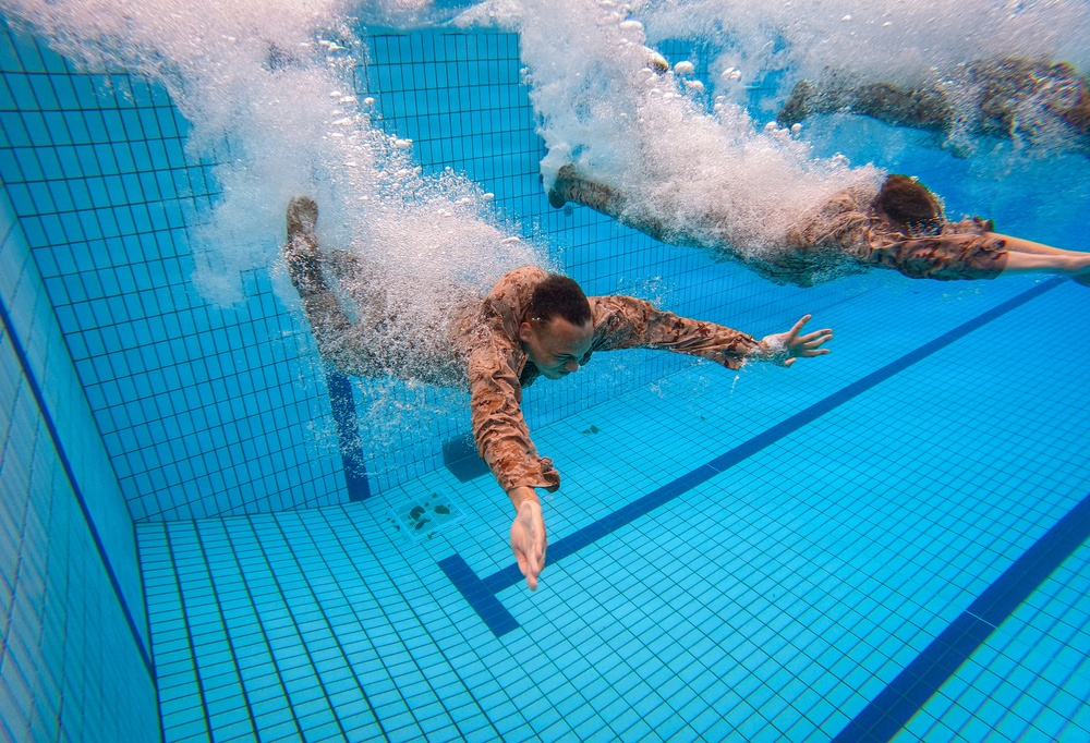 An underwater photograph captures two individuals wearing military camouflage uniforms diving or jumping into a swimming pool. The image is taken from below the water surface, showing the swimmers mid-entry with large air bubble trails surrounding them. The tiled pool floor and lane markers are clearly visible beneath the swimmers.