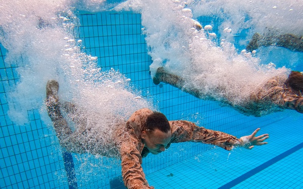 An underwater photograph captures two individuals wearing military camouflage uniforms diving or jumping into a swimming pool. The image is taken from below the water surface, showing the swimmers mid-entry with large air bubble trails surrounding them. The tiled pool floor and lane markers are clearly visible beneath the swimmers.