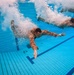An underwater photograph captures two individuals wearing military camouflage uniforms diving or jumping into a swimming pool. The image is taken from below the water surface, showing the swimmers mid-entry with large air bubble trails surrounding them. The tiled pool floor and lane markers are clearly visible beneath the swimmers.
