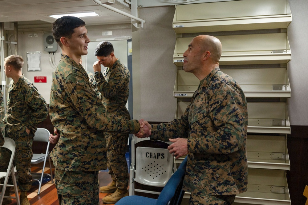 Two U.S. Marines in MARPAT camouflage uniforms are shaking hands in what appears to be an indoor military facility, possibly aboard a naval vessel. A chair labeled 'CHAPS' is visible in the foreground, suggesting a chaplain's area or religious services space. Additional Marines are visible in the background.