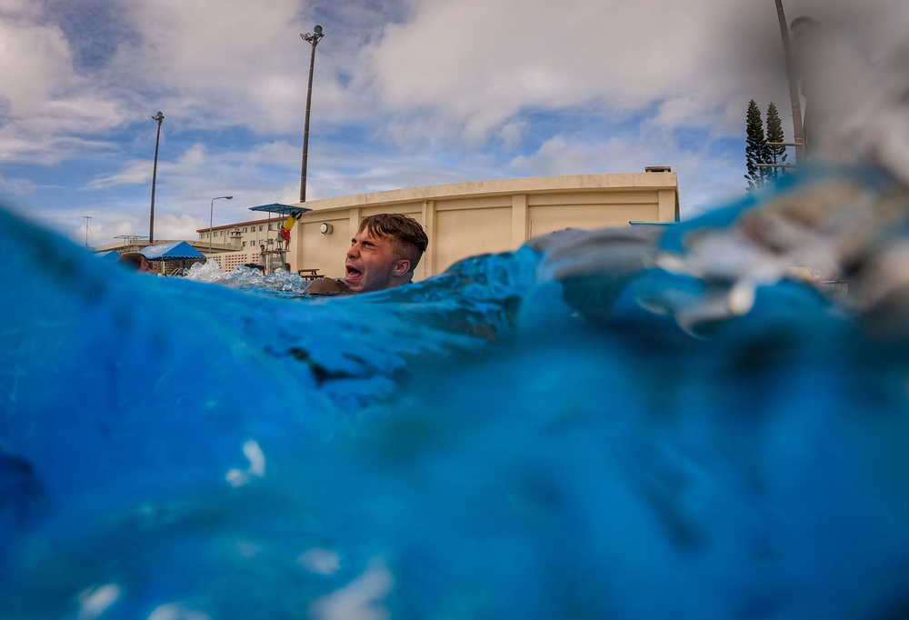 A low water-level perspective photograph captures a person emerging from or swimming in a pool, with their head just above the water surface and mouth open. The dramatic camera angle is positioned at water level, making the blue water dominate the foreground. The setting appears to be an outdoor pool facility with utility poles, a building, and trees visible in the background under a partly cloudy sky.