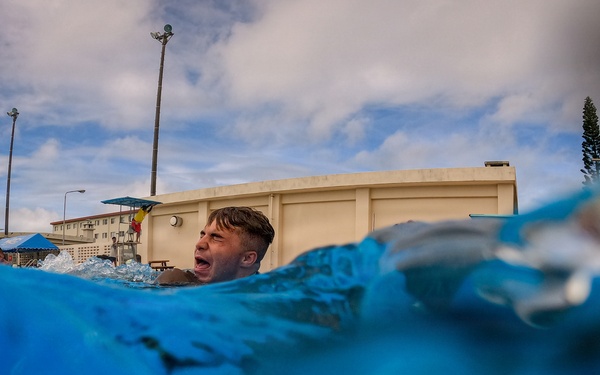 A low water-level perspective photograph captures a person emerging from or swimming in a pool, with their head just above the water surface and mouth open. The dramatic camera angle is positioned at water level, making the blue water dominate the foreground. The setting appears to be an outdoor pool facility with utility poles, a building, and trees visible in the background under a partly cloudy sky.
