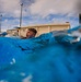 A low water-level perspective photograph captures a person emerging from or swimming in a pool, with their head just above the water surface and mouth open. The dramatic camera angle is positioned at water level, making the blue water dominate the foreground. The setting appears to be an outdoor pool facility with utility poles, a building, and trees visible in the background under a partly cloudy sky.