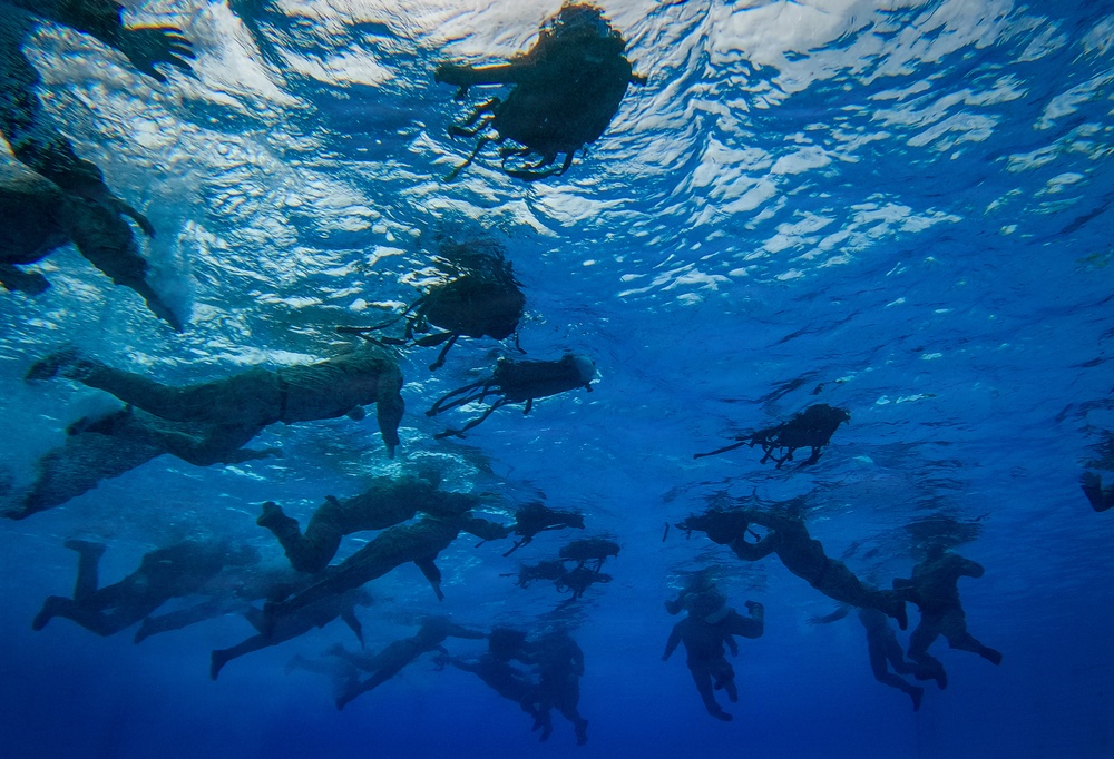 An underwater photograph taken from below the surface showing a large group of swimmers or divers in open blue water. The image is captured looking upward toward the water surface, revealing multiple human silhouettes swimming at or just below the surface. The deep blue water and rippling surface light create a dramatic visual effect.