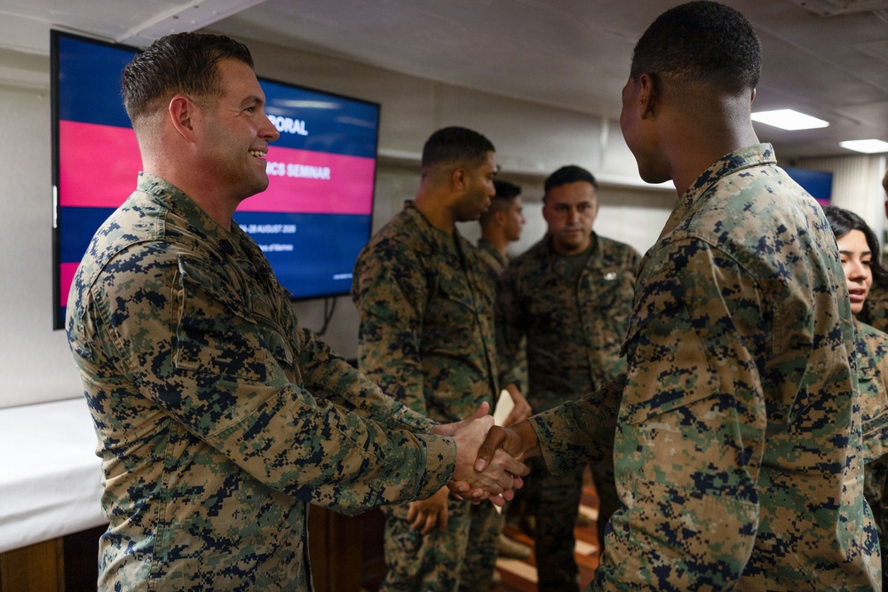 Military personnel in U.S. Marine Corps MARPAT camouflage uniforms are gathered in an indoor setting, with two individuals shaking hands in the foreground. A presentation screen in the background displays partial text including what appears to be 'Seminar' and a date referencing August, suggesting a formal military educational or briefing event. The atmosphere appears professional and cordial.
