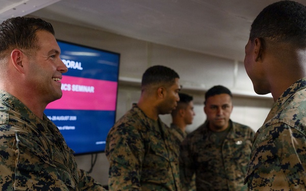 Military personnel in U.S. Marine Corps MARPAT camouflage uniforms are gathered in an indoor setting, with two individuals shaking hands in the foreground. A presentation screen in the background displays partial text including what appears to be 'Seminar' and a date referencing August, suggesting a formal military educational or briefing event. The atmosphere appears professional and cordial.