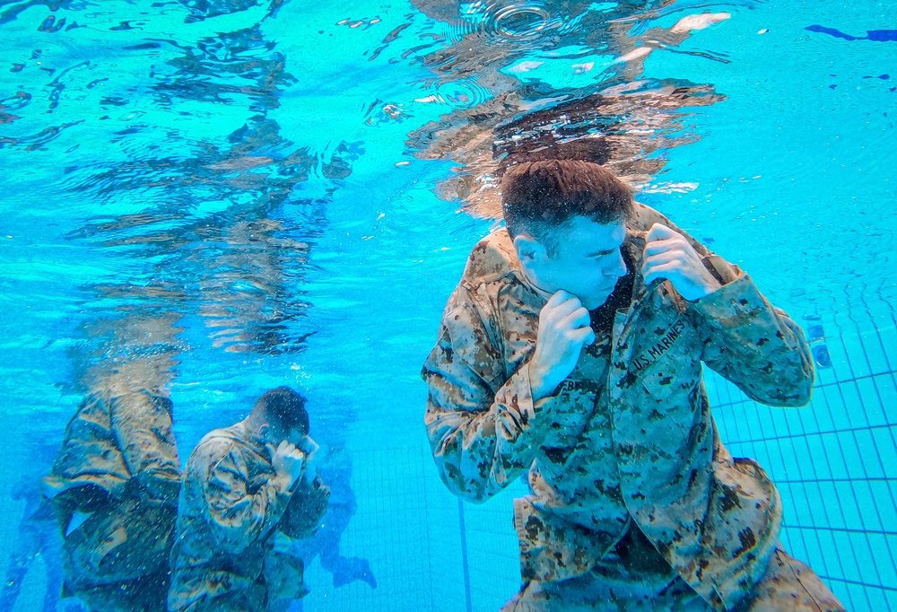 An underwater photograph captures two U.S. Marines in full camouflage uniforms submerged in a swimming pool. Both individuals are in a defensive boxing stance with fists raised, suggesting a water survival or combat training exercise. The image is taken from an underwater perspective, showing the pool's tiled bottom and the water surface above.
