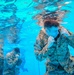 An underwater photograph captures two U.S. Marines in full camouflage uniforms submerged in a swimming pool. Both individuals are in a defensive boxing stance with fists raised, suggesting a water survival or combat training exercise. The image is taken from an underwater perspective, showing the pool's tiled bottom and the water surface above.