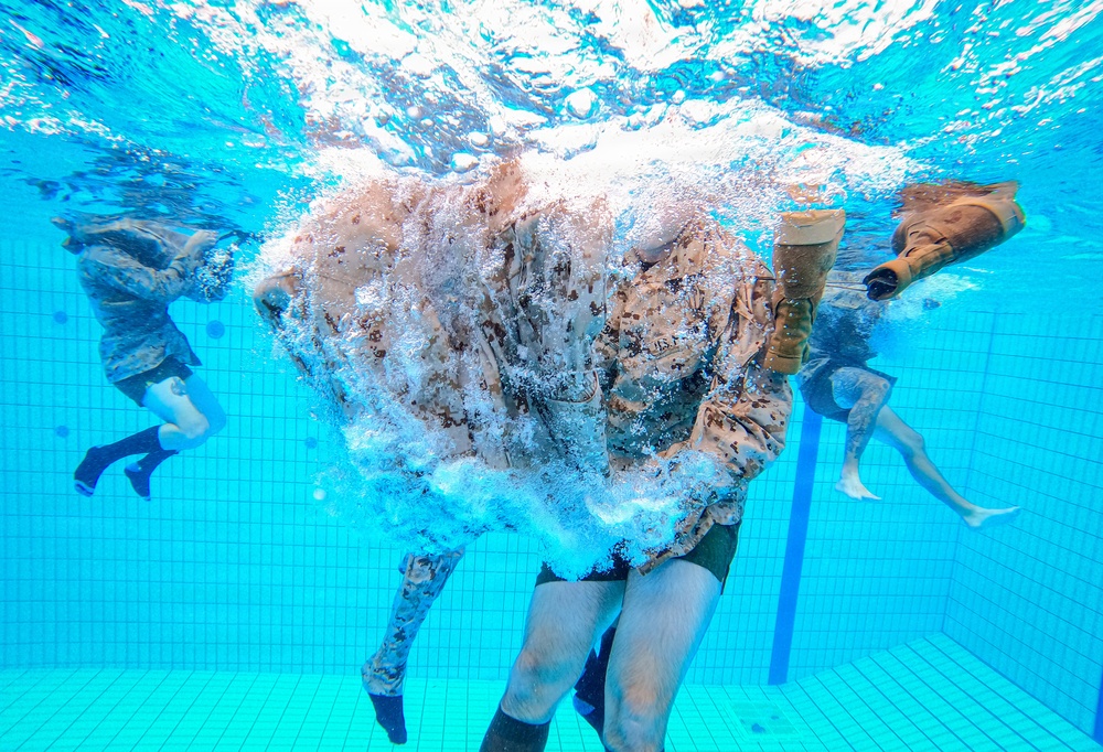 An underwater photograph taken inside a swimming pool showing multiple individuals in military camouflage uniforms submerged in water. The central figure appears to have just entered the water, surrounded by a large burst of air bubbles. This appears to be a military water survival or combat swimmer training exercise.