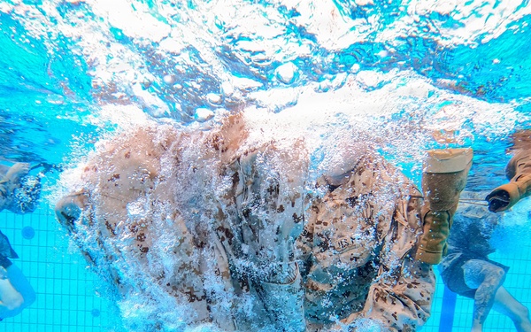 An underwater photograph taken inside a swimming pool showing multiple individuals in military camouflage uniforms submerged in water. The central figure appears to have just entered the water, surrounded by a large burst of air bubbles. This appears to be a military water survival or combat swimmer training exercise.