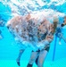 An underwater photograph taken inside a swimming pool showing multiple individuals in military camouflage uniforms submerged in water. The central figure appears to have just entered the water, surrounded by a large burst of air bubbles. This appears to be a military water survival or combat swimmer training exercise.