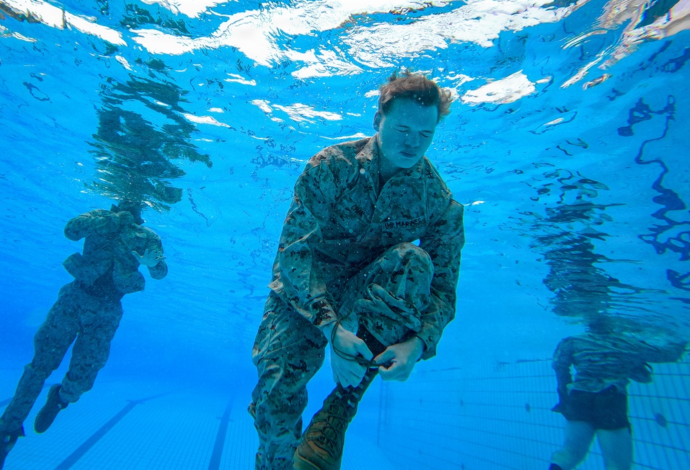 An underwater photograph taken inside a swimming pool showing U.S. Marines in full camouflage uniforms (MARPAT pattern) submerged and performing what appears to be a water survival or combat swimmer training exercise. The primary subject in the foreground appears to be manipulating equipment or clothing while underwater. At least two additional personnel are visible in the background.