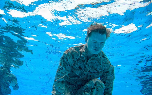 An underwater photograph taken inside a swimming pool showing U.S. Marines in full camouflage uniforms (MARPAT pattern) submerged and performing what appears to be a water survival or combat swimmer training exercise. The primary subject in the foreground appears to be manipulating equipment or clothing while underwater. At least two additional personnel are visible in the background.