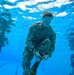 An underwater photograph taken inside a swimming pool showing U.S. Marines in full camouflage uniforms (MARPAT pattern) submerged and performing what appears to be a water survival or combat swimmer training exercise. The primary subject in the foreground appears to be manipulating equipment or clothing while underwater. At least two additional personnel are visible in the background.