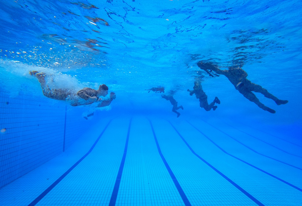 An underwater photograph taken inside an indoor swimming pool showing multiple people submerged at various depths and distances. The image is captured from a low angle near the pool floor, revealing lane markings on the bottom and the water surface above. The swimmers appear to be wearing clothing or uniforms rather than typical swimwear, suggesting a training or military exercise.