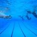 An underwater photograph taken inside an indoor swimming pool showing multiple people submerged at various depths and distances. The image is captured from a low angle near the pool floor, revealing lane markings on the bottom and the water surface above. The swimmers appear to be wearing clothing or uniforms rather than typical swimwear, suggesting a training or military exercise.