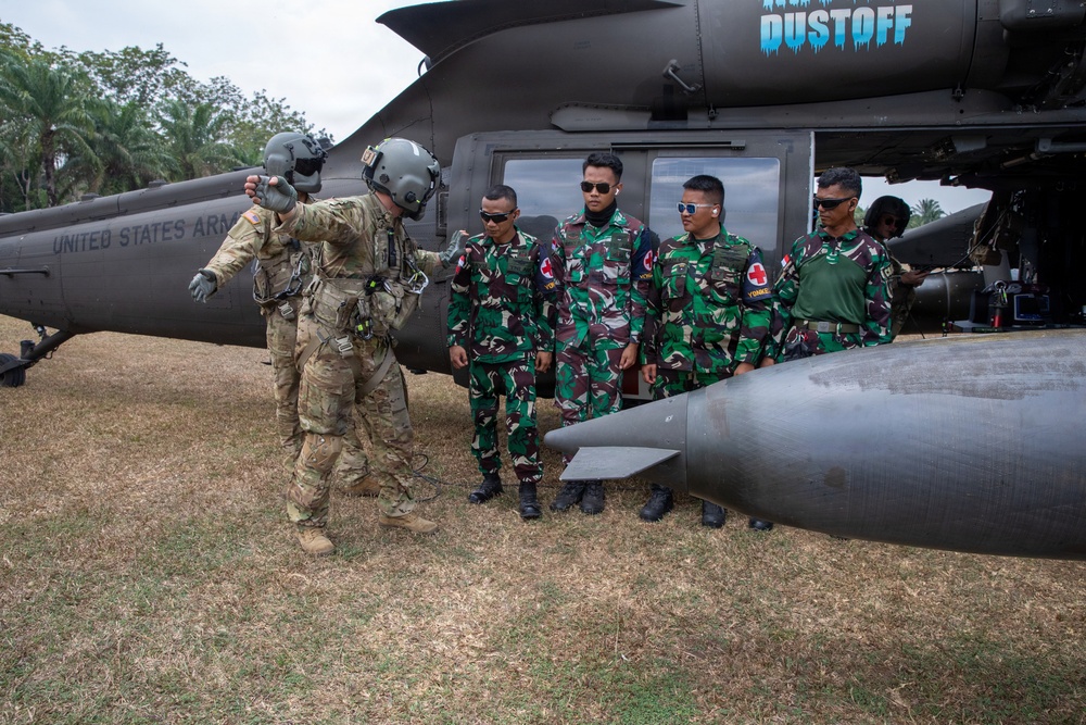 A United States Army aviator in flight gear is briefing or instructing a group of foreign military personnel wearing camouflage uniforms with red cross medical patches near a UH-60 Black Hawk helicopter marked 'DUSTOFF'. The helicopter bears 'United States Army' markings, indicating this is a medical evacuation (MEDEVAC) aircraft. This appears to be a joint military training or familiarization exercise.