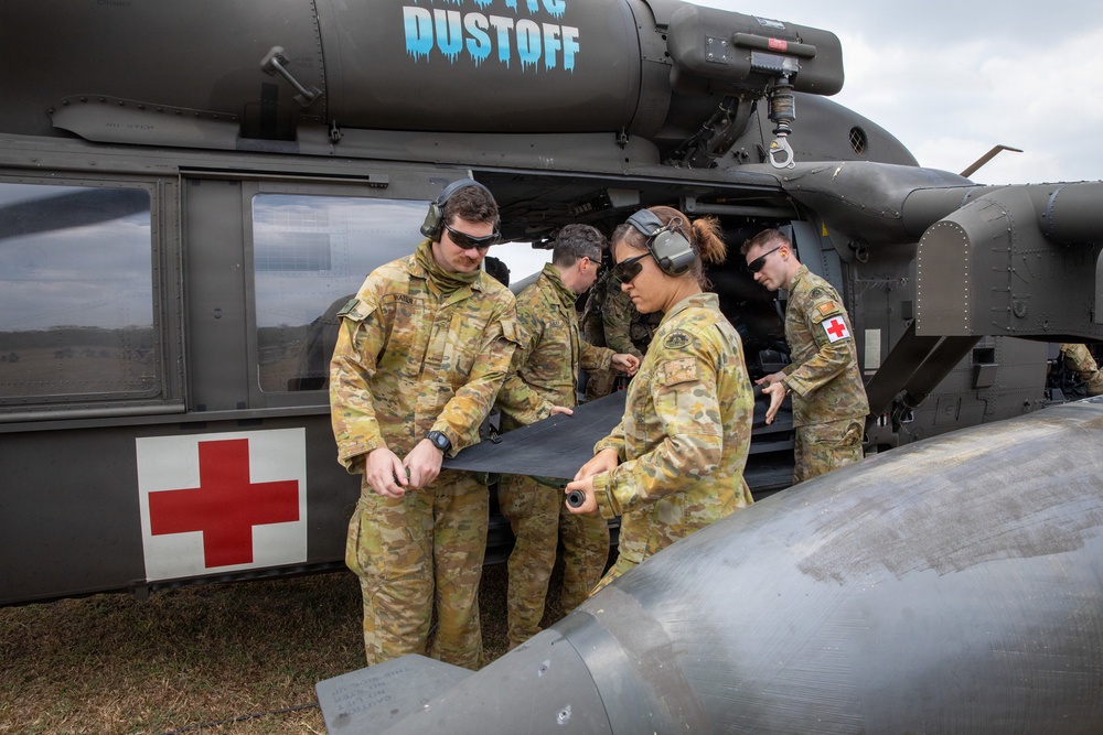 A group of military personnel in Australian Defence Force (ADF) multicam camouflage uniforms are working alongside a medical evacuation helicopter marked with a Red Cross symbol and the word 'DUSTOFF'. The crew members appear to be loading or preparing a stretcher/litter for a MEDEVAC operation. The helicopter is a UH-60 Black Hawk variant configured for aeromedical evacuation.