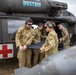 A group of military personnel in Australian Defence Force (ADF) multicam camouflage uniforms are working alongside a medical evacuation helicopter marked with a Red Cross symbol and the word 'DUSTOFF'. The crew members appear to be loading or preparing a stretcher/litter for a MEDEVAC operation. The helicopter is a UH-60 Black Hawk variant configured for aeromedical evacuation.
