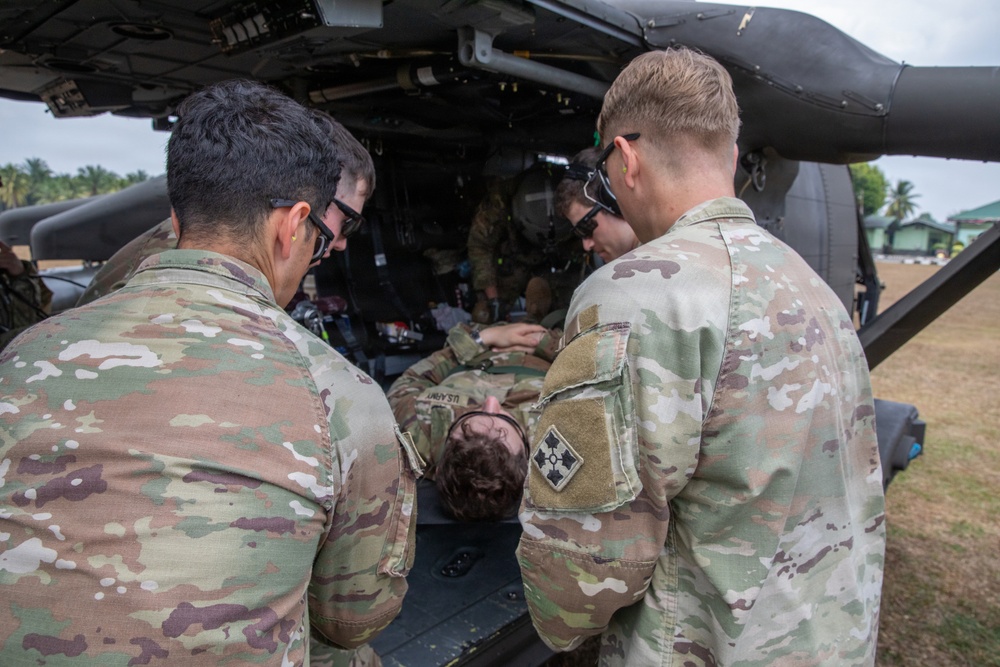 Military personnel in OCP (Operational Camouflage Pattern) uniforms are conducting a medical evacuation (MEDEVAC) exercise or operation near a military helicopter. A simulated or actual casualty in uniform is being loaded or attended to at the rear of the aircraft. Multiple soldiers are actively engaged in the patient transfer process.
