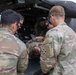 Military personnel in OCP (Operational Camouflage Pattern) uniforms are conducting a medical evacuation (MEDEVAC) exercise or operation near a military helicopter. A simulated or actual casualty in uniform is being loaded or attended to at the rear of the aircraft. Multiple soldiers are actively engaged in the patient transfer process.