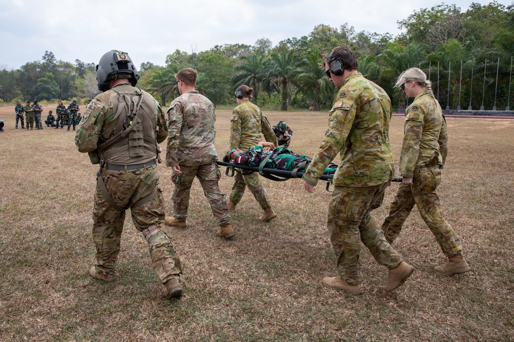 Military personnel in camouflage uniforms are carrying a person on a stretcher across an open grassy field during what appears to be a joint military training exercise. The group includes both male and female soldiers wearing hearing protection, with one individual wearing a flight helmet. A separate group of uniformed personnel can be seen observing in the background.