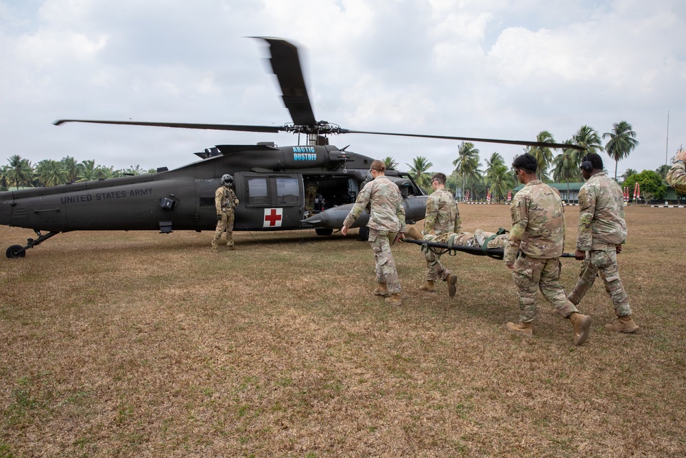 U.S. Army soldiers are conducting a medical evacuation (MEDEVAC) exercise, carrying a patient on a litter toward a UH-60 Black Hawk helicopter marked with a red cross. The helicopter is labeled 'United States Army' and bears the designation 'Arctic Dustoff,' indicating a medical evacuation unit. The scene appears to take place at a tropical location, likely during a joint military exercise.