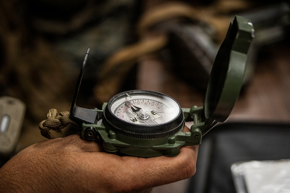 A hand is holding an open military-style lensatic compass in olive drab/green casing. The compass face is clearly visible with cardinal directions (W and E visible) and degree markings in both black and red. The background is blurred, showing what appears to be military or outdoor gear.