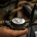 A hand is holding an open military-style lensatic compass in olive drab/green casing. The compass face is clearly visible with cardinal directions (W and E visible) and degree markings in both black and red. The background is blurred, showing what appears to be military or outdoor gear.