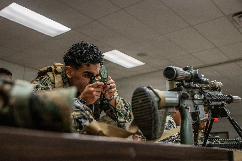 A U.S. Marine in woodland MARPAT camouflage uniform and tactical vest is looking through what appears to be a military lensatic compass or laser rangefinder in an indoor classroom or training facility. Additional Marines are visible in the background, and a scoped rifle on a bipod is positioned on the table to the right. The setting appears to be a formal military training or instruction environment.