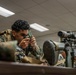 A U.S. Marine in woodland MARPAT camouflage uniform and tactical vest is looking through what appears to be a military lensatic compass or laser rangefinder in an indoor classroom or training facility. Additional Marines are visible in the background, and a scoped rifle on a bipod is positioned on the table to the right. The setting appears to be a formal military training or instruction environment.