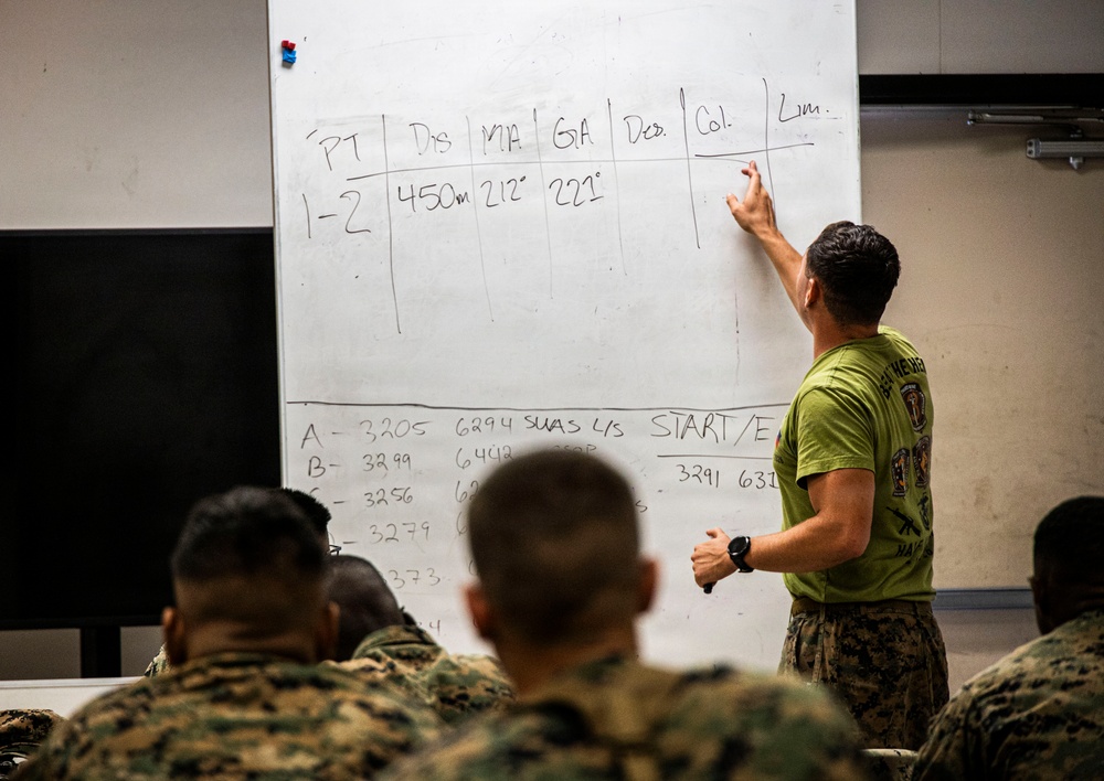 A military instructor in a green t-shirt and camouflage trousers is writing on a large whiteboard during a classroom training session. The whiteboard contains a table with columns labeled PT, Dis, MA, GA, Des, Col, and Lim, with data entries including distances and degree measurements. Several military personnel in camouflage uniforms are seated facing the instructor, taking notes or observing.