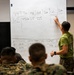 A military instructor in a green t-shirt and camouflage trousers is writing on a large whiteboard during a classroom training session. The whiteboard contains a table with columns labeled PT, Dis, MA, GA, Des, Col, and Lim, with data entries including distances and degree measurements. Several military personnel in camouflage uniforms are seated facing the instructor, taking notes or observing.
