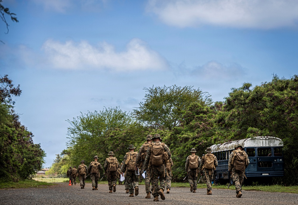 A group of military personnel in camouflage uniforms and tactical gear are marching along a paved road in a lush, green environment. They are carrying backpacks and appear to be on a training exercise or patrol. An old, weathered blue bus is parked on the right side of the road near dense vegetation.