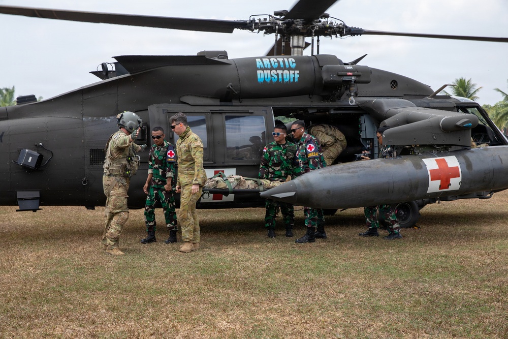 Military medical personnel from what appear to be U.S. and Indonesian armed forces are conducting a MEDEVAC exercise beside a black UH-60 Black Hawk helicopter marked 'Arctic Dustoff'. Personnel are carrying a simulated casualty on a stretcher toward the helicopter. The helicopter bears a red cross medical insignia on its external fuel tank, indicating its medical evacuation role.
